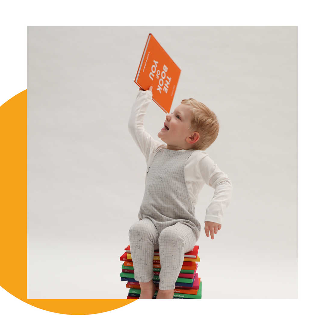 Toddler holding childhood memory journal in the air sat on top of a pile of memory journals in different colours